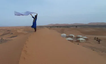 bedouin man holding white scarf in wind atop dune with nomad tents and big dunes in background