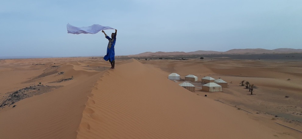 bedouin man holding white scarf in wind atop dune with nomad tents and big dunes in background